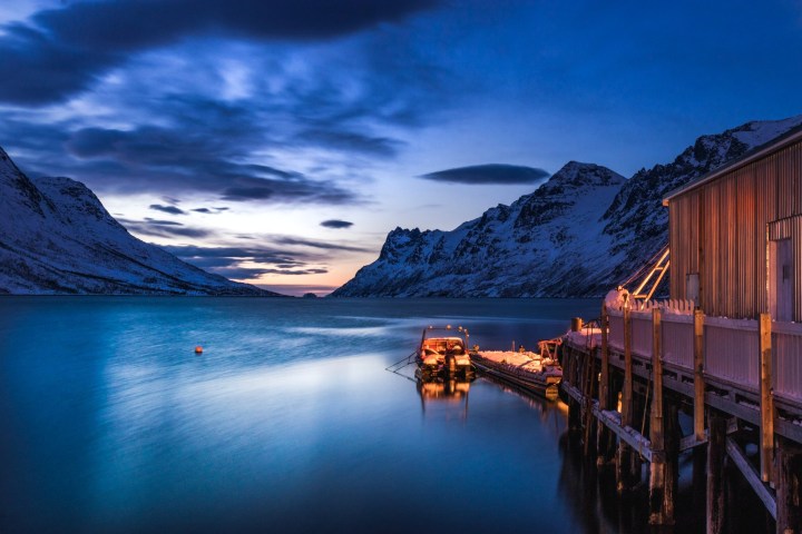 Scenic view of a dock and cabin by a calm fjord with snow-capped mountains at twilight.