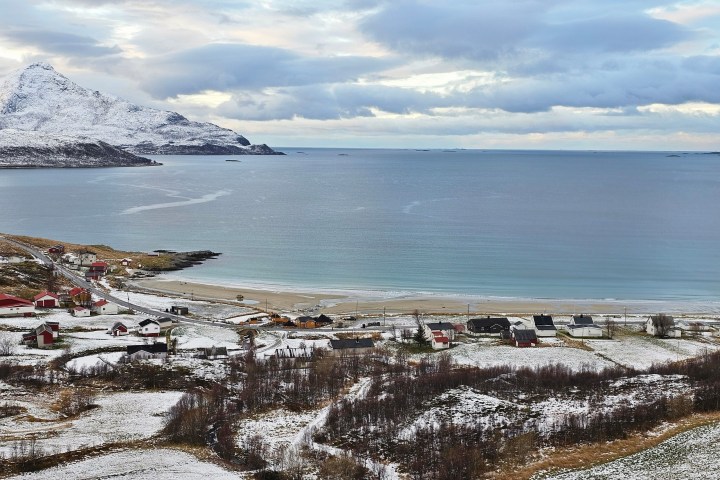 Snow-dusted coastal landscape with mountains, sea, and scattered houses under a cloudy sky.