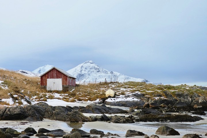 Red cabin on snowy coastal landscape with mountains in background.