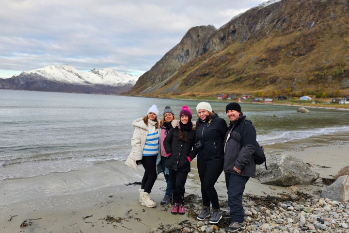 Group of people in winter clothing on a rocky beach by mountains.