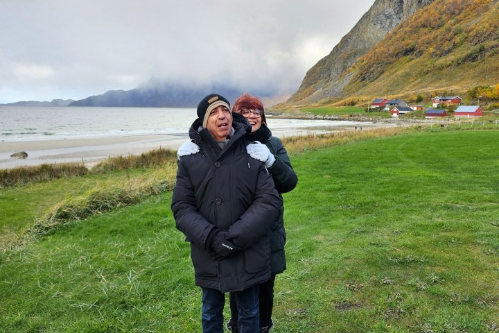 Two people in winter coats standing on a grassy area near mountains and sea.