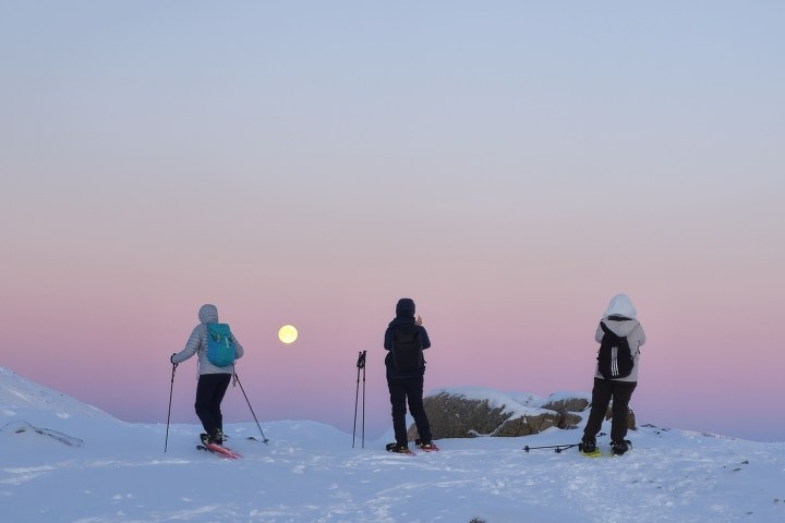Three skiers on snowy hill at sunset with a visible full moon.