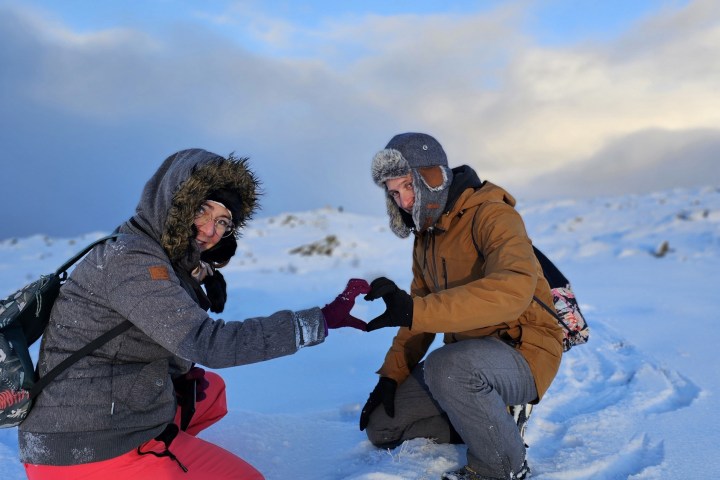 Two people in winter gear kneeling in snow, making heart shape with hands.