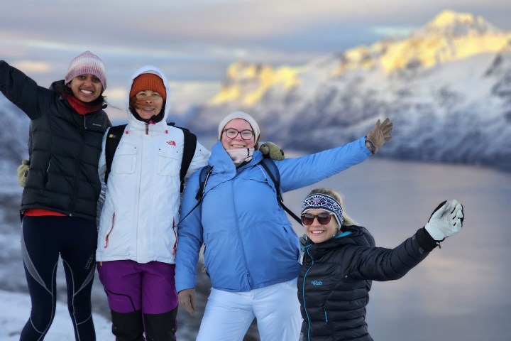 Four people in winter attire smiling, posing with raised arms by a snowy mountain lake.