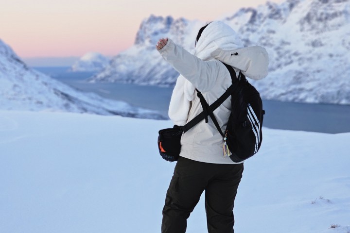 Person in snow gear faces mountains, standing in snow with arms raised.